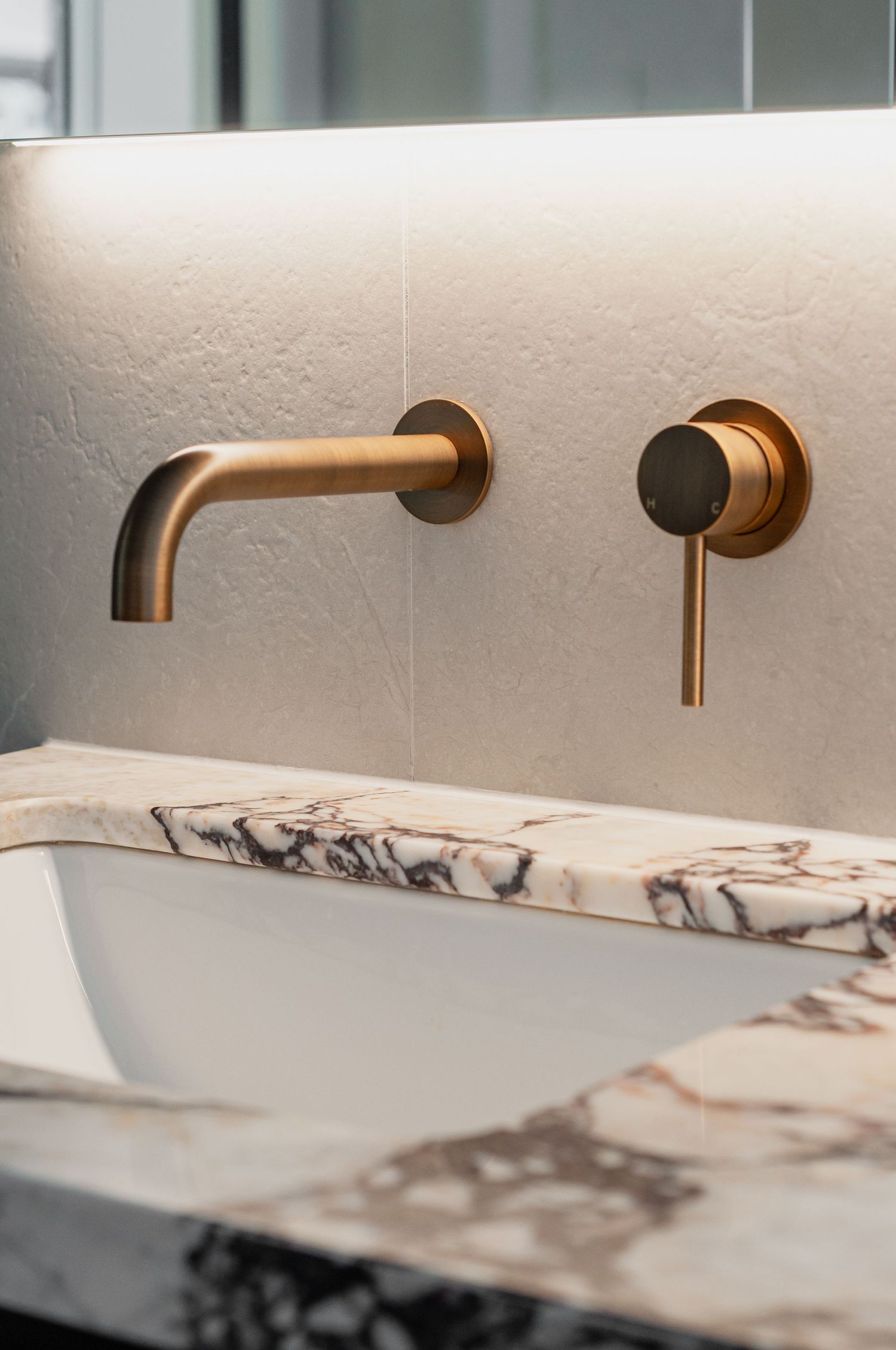 Close-up of brass wall-mounted faucet and tap against white textured wall with marble counter.