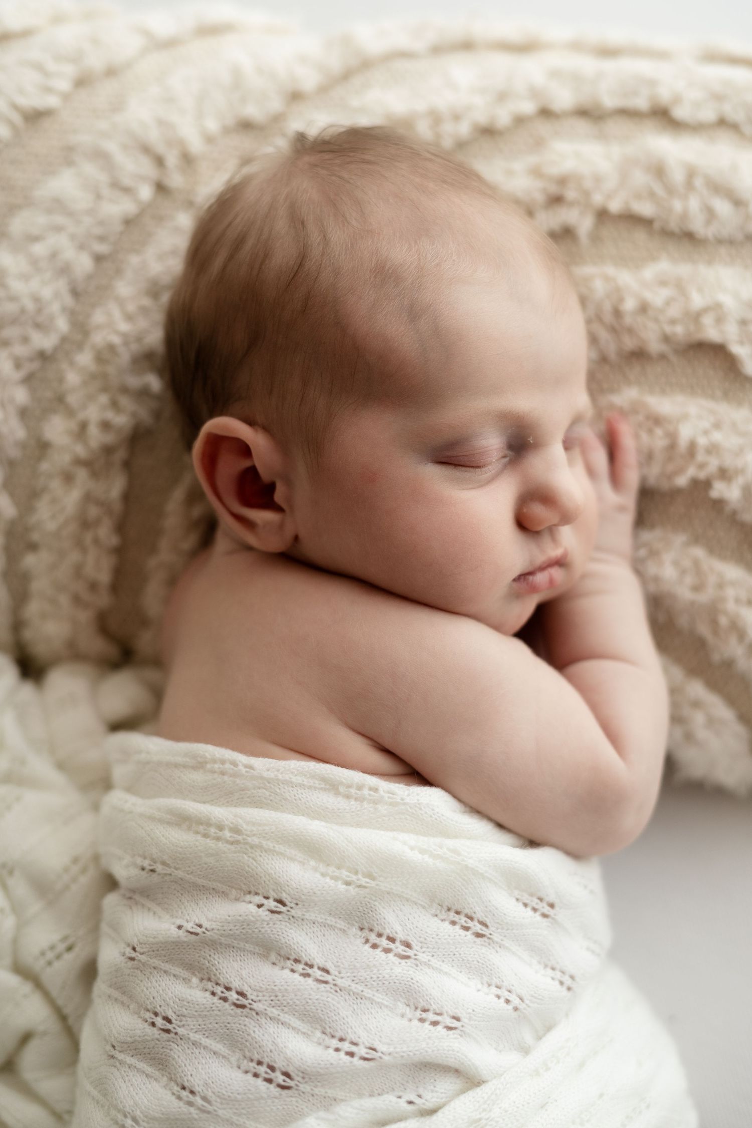 Newborn baby sleeping peacefully wrapped in white knit blanket on fluffy cream surface.
