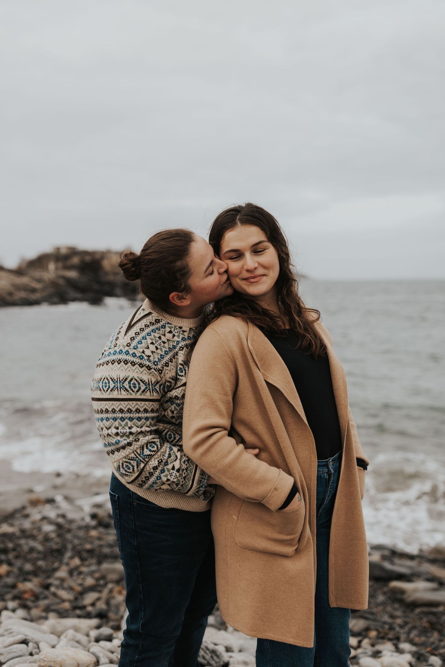 A romantic moment between two people wearing cozy sweaters on a rocky coastline on an overcast day.
