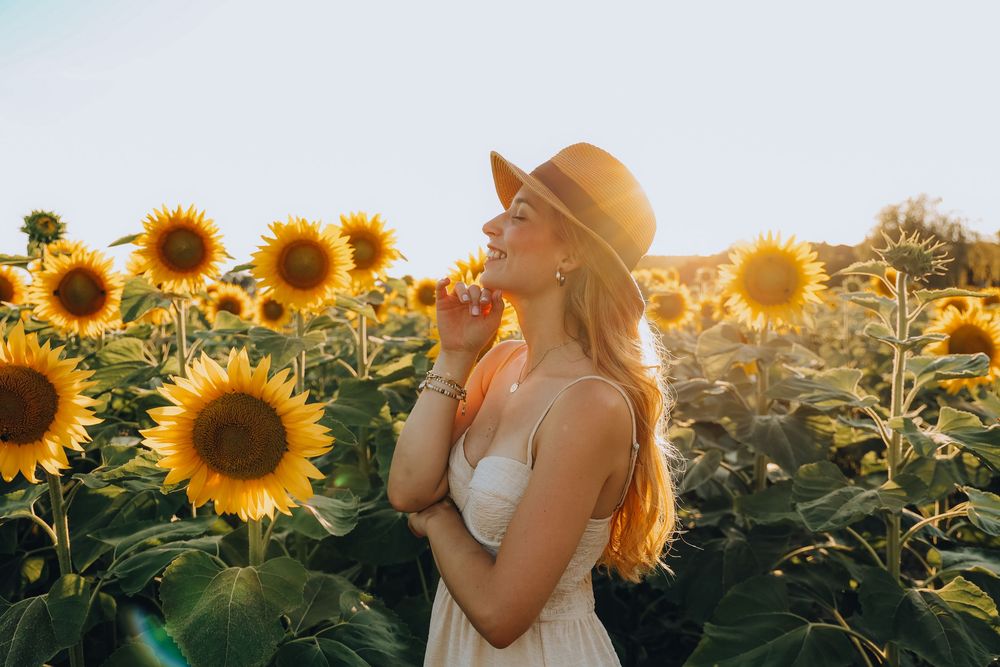 Serene woman in hat and white dress enjoying sunflower field in warm sunlight.