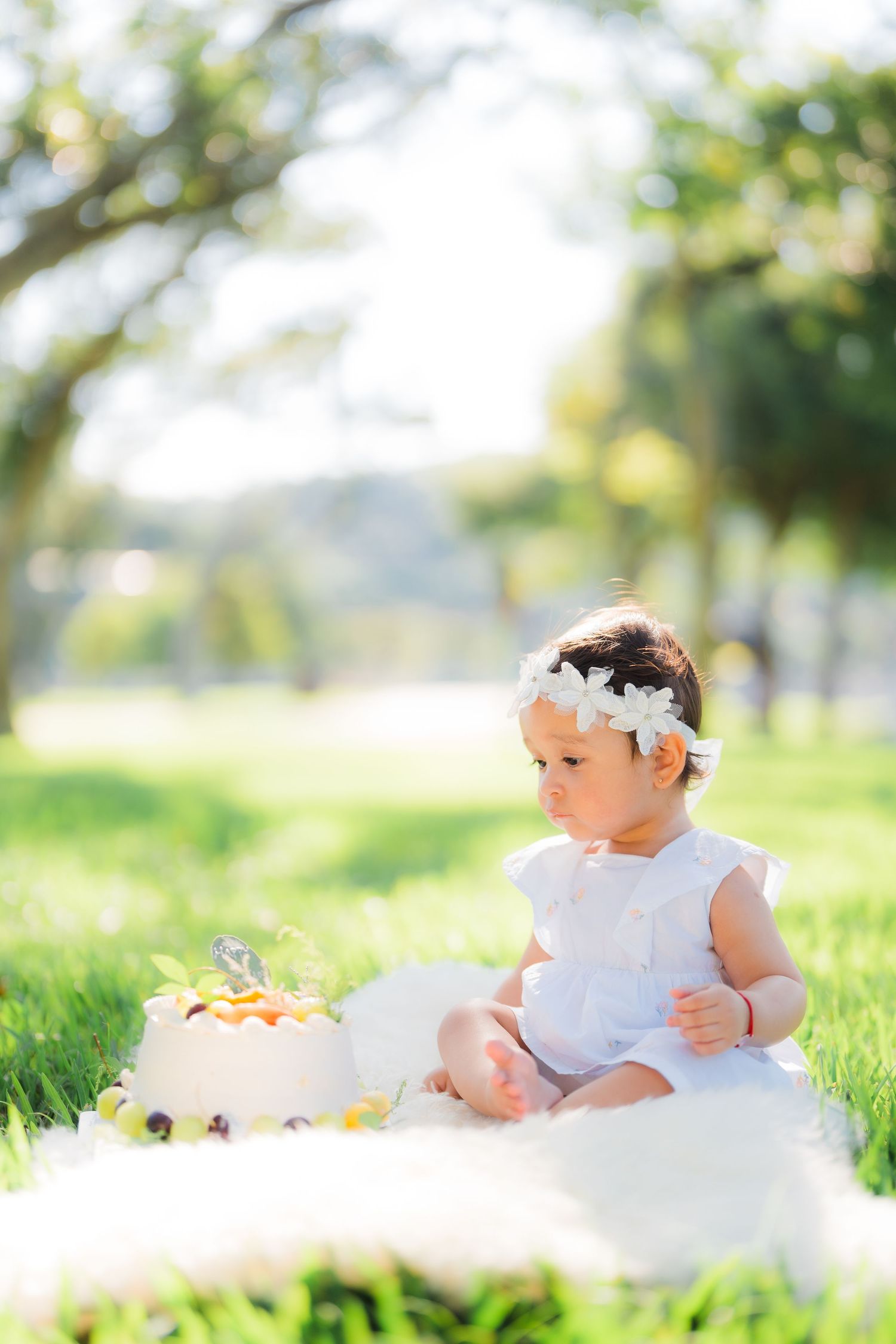 Baby in white dress sits on blanket next to birthday cake in sunny outdoor setting.