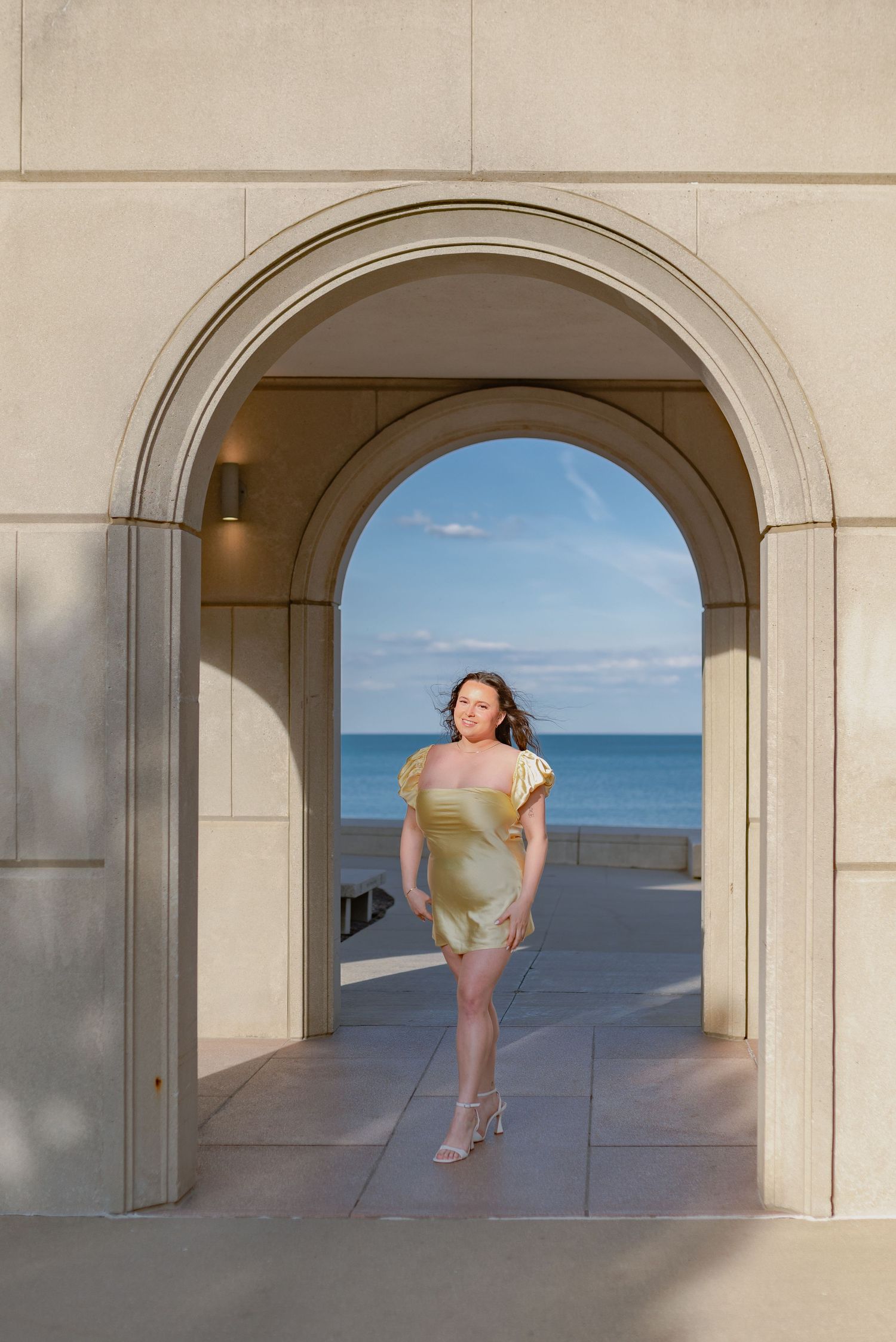 Figure in a yellow dress poses beneath an elegant archway overlooking the ocean on a sunny day.