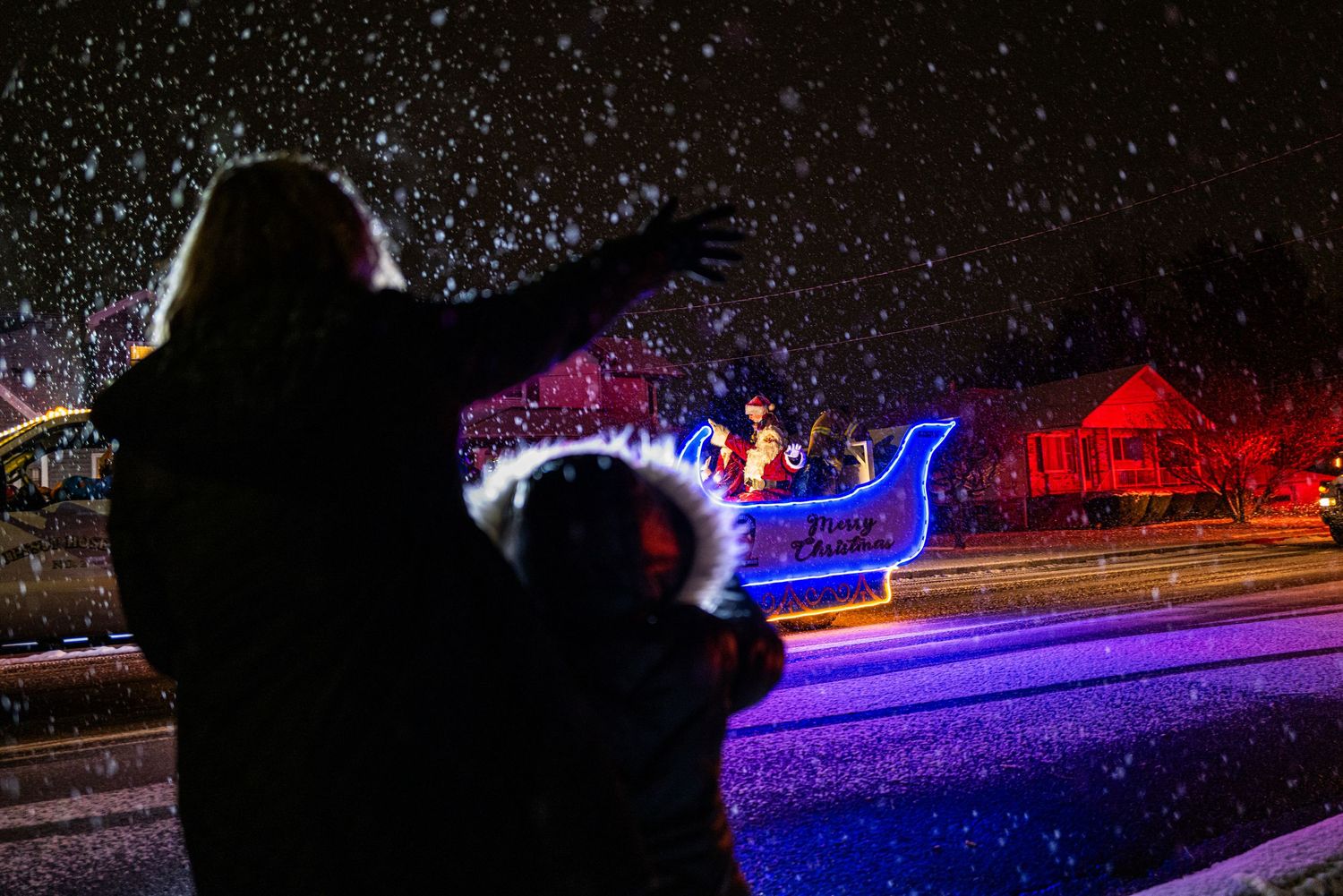 Colorful holiday light display illuminating a path through trees and buildings at night.