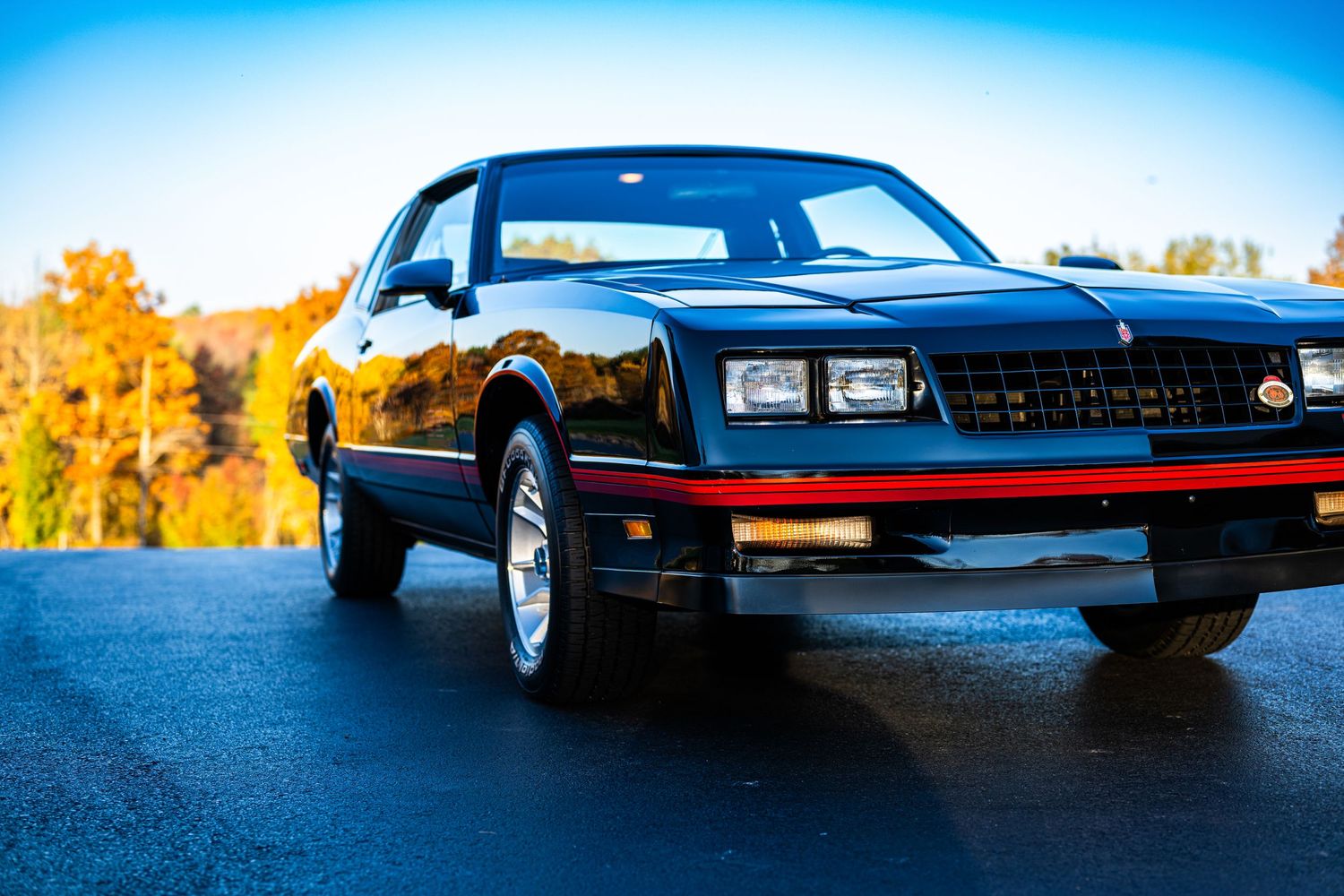 Low-angle panoramic view of a classic black sports car against a blue sky and autumn foliage.