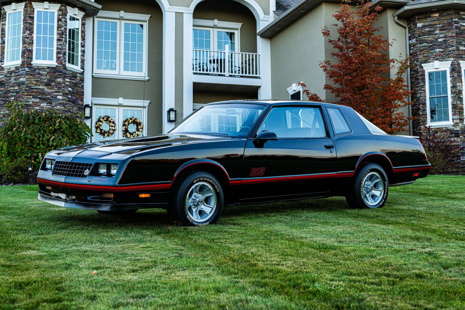 Black vintage car parked on grass in front of large suburban houses with fall colors.