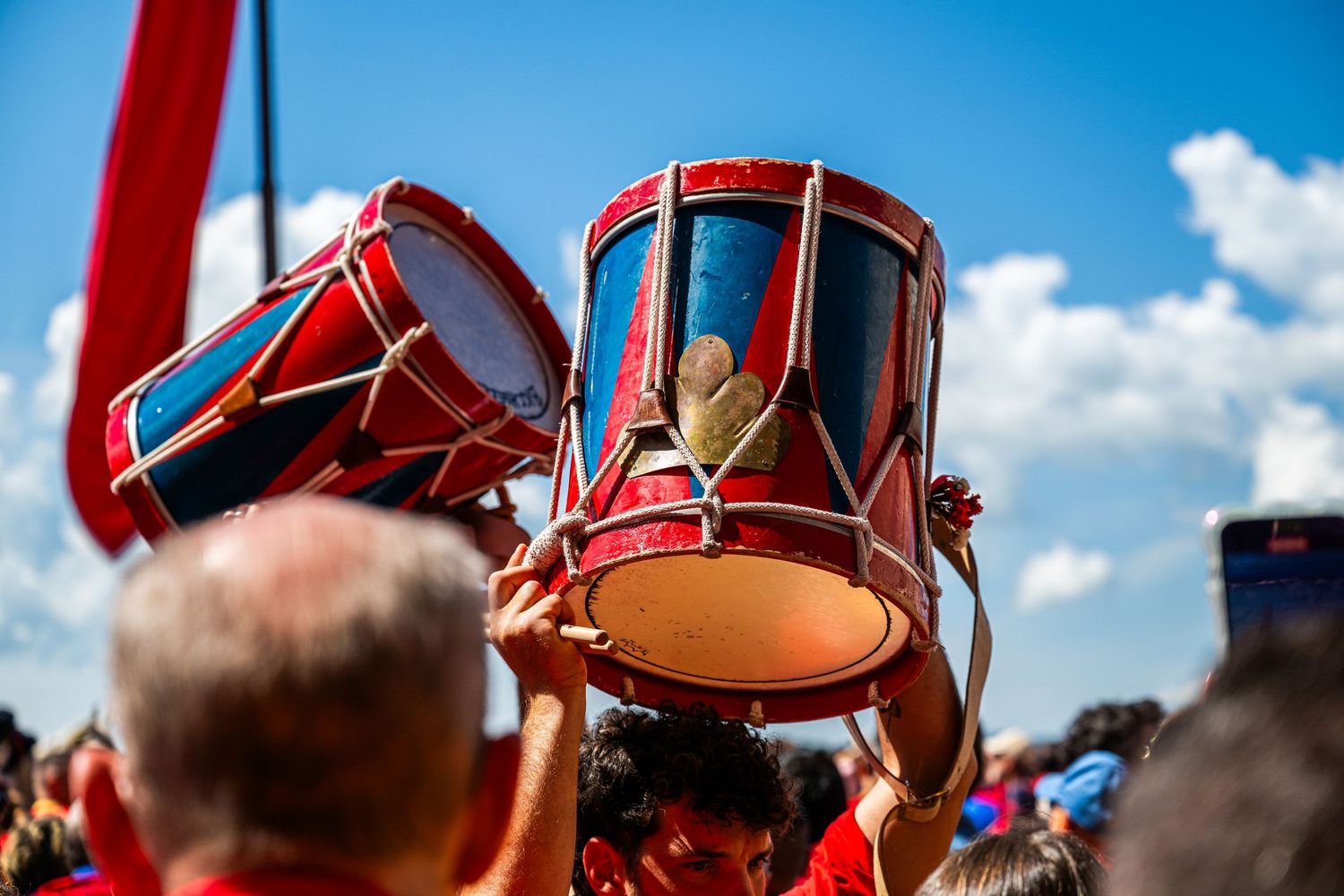 Red decorative drums held aloft at a vibrant outdoor festival parade under a bright blue sky.