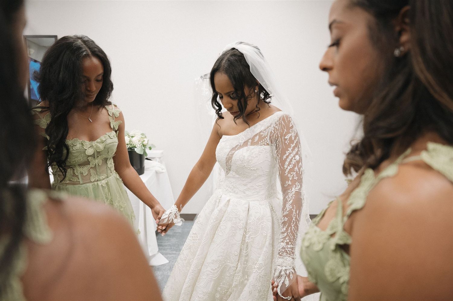 People gather around a person wearing an elegant white lace wedding gown with long sleeves in a bridal suite.
