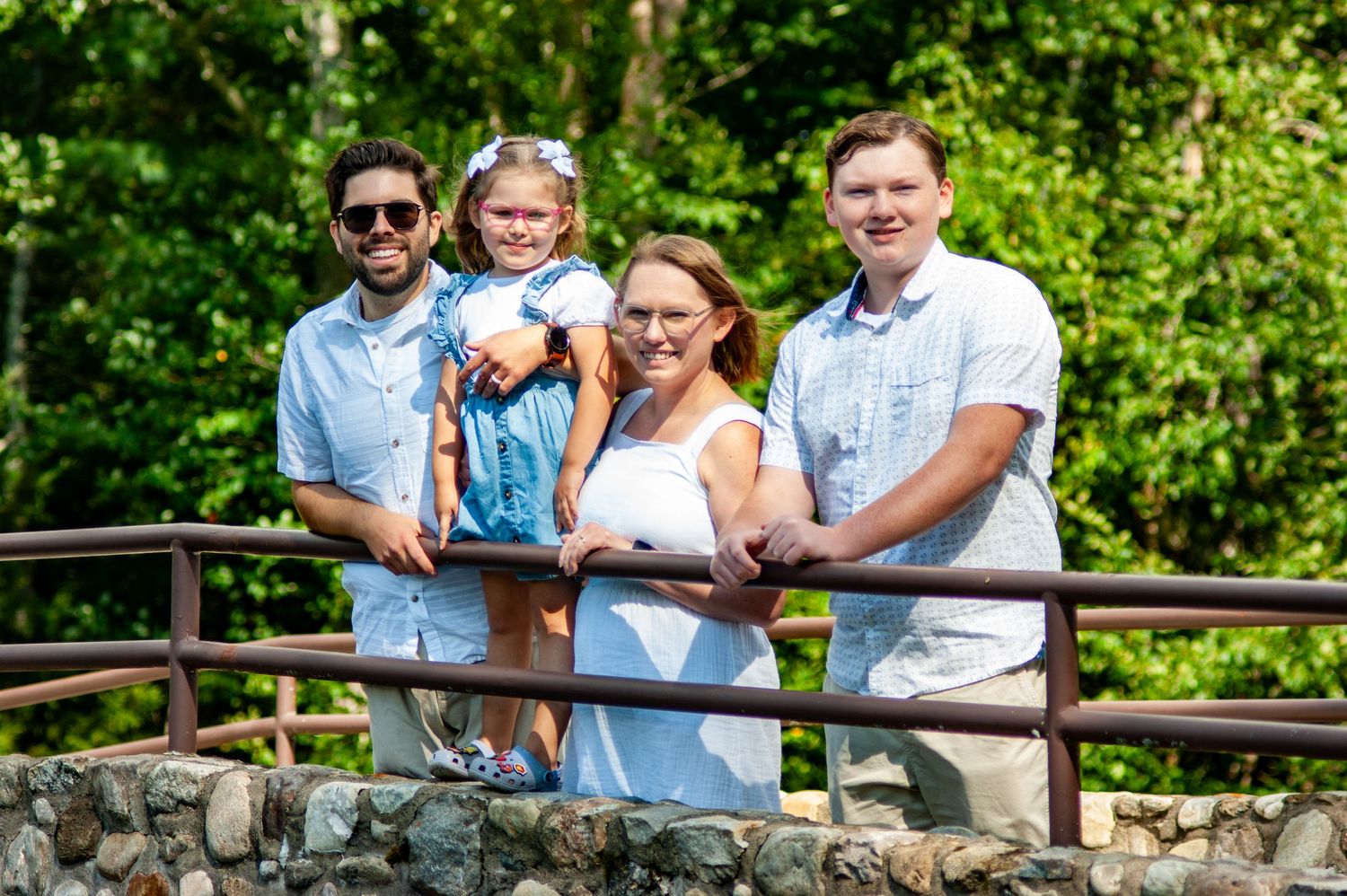 A family poses together on a stone bridge surrounded by lush green forest on a bright summer day.