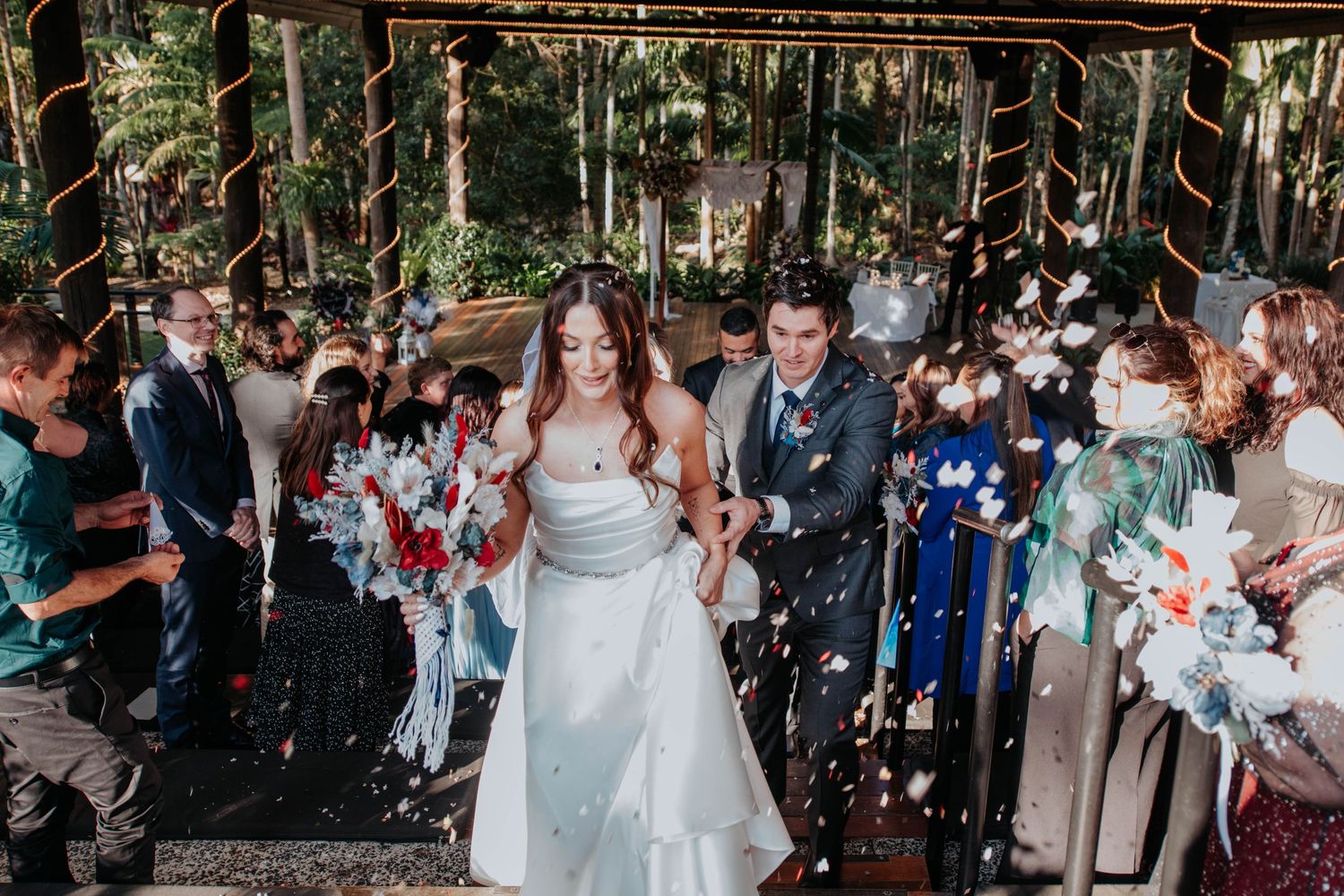Wedding couple exits ceremony under shower of confetti in outdoor forest setting with guests in attendance.
