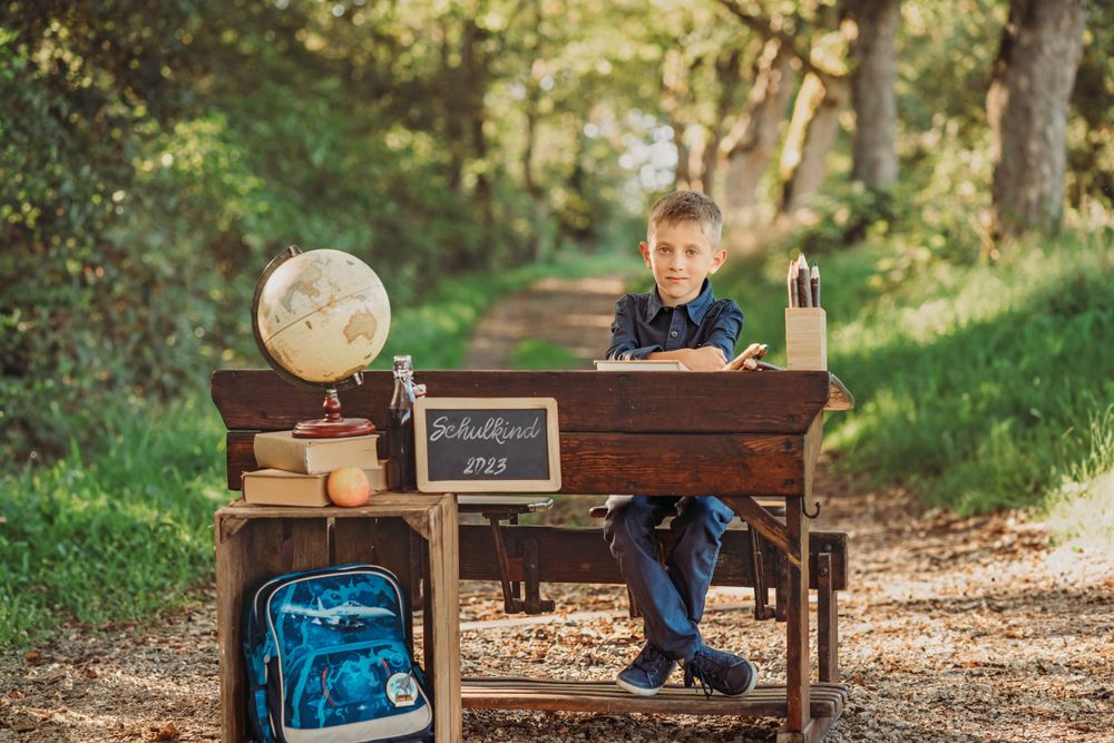Student sits at vintage desk with globe and backpack in wooded area for back to school photo.