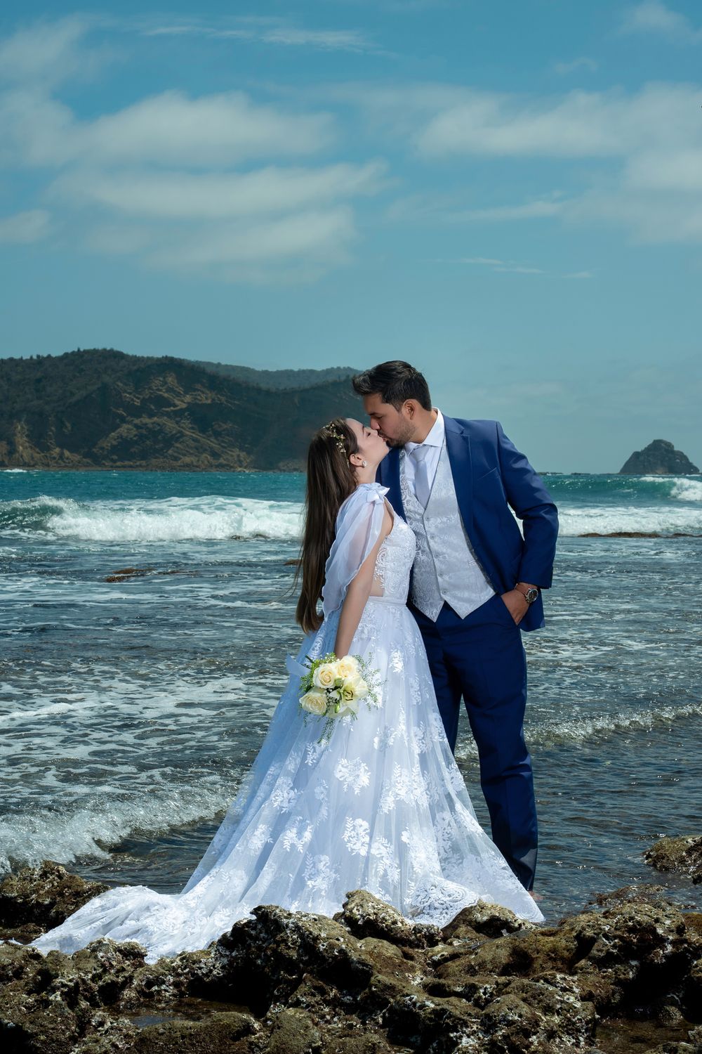 Romantic coastal wedding photoshoot with crashing waves and rocky shoreline in the background.