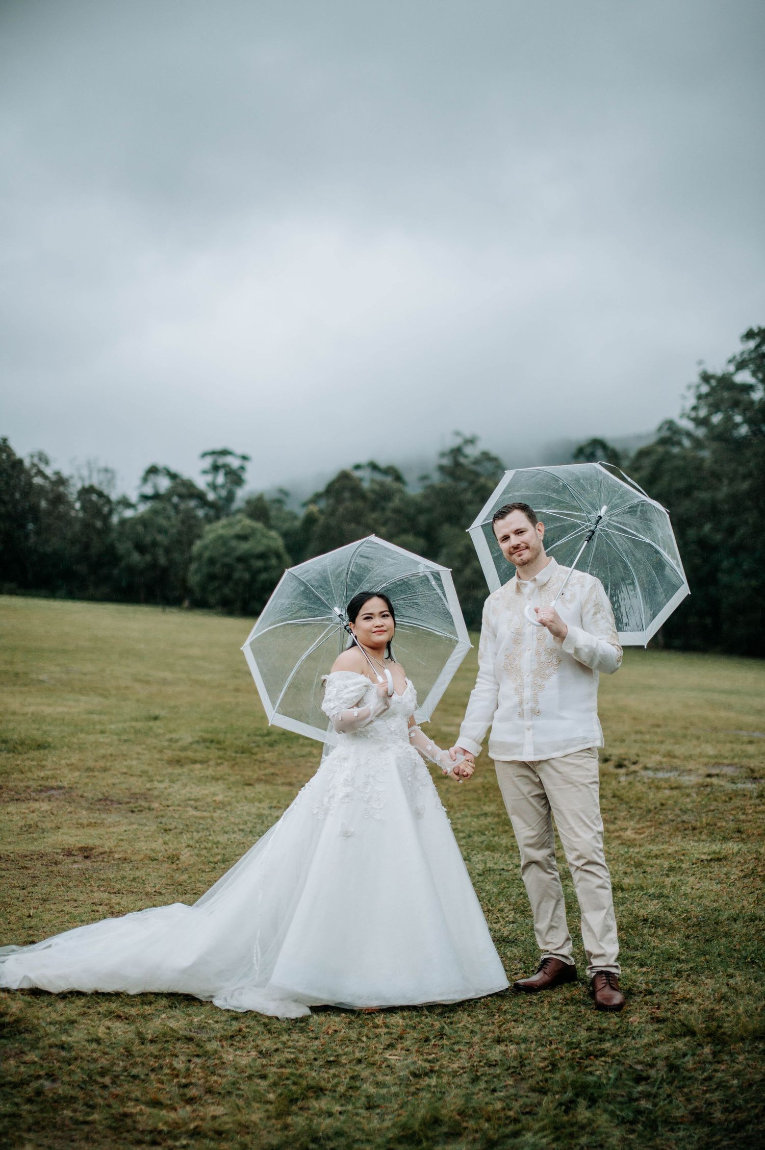 Wedding couple poses with clear umbrellas on overcast day in countryside.