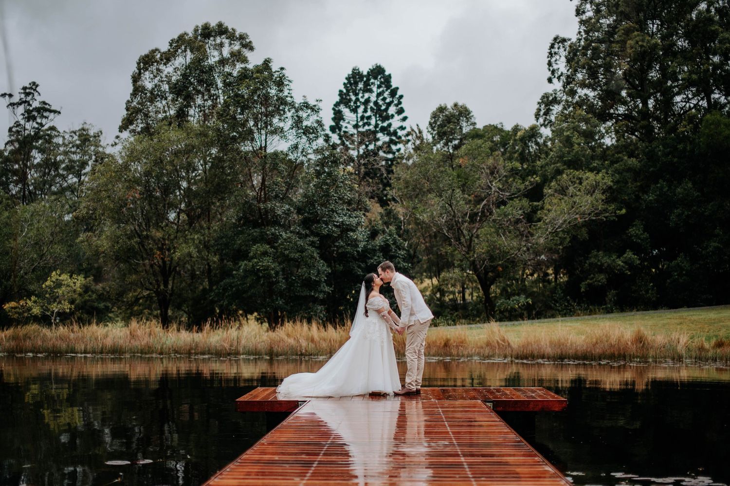 A couple embraces on a wooden dock over a serene lake surrounded by trees at sunset.