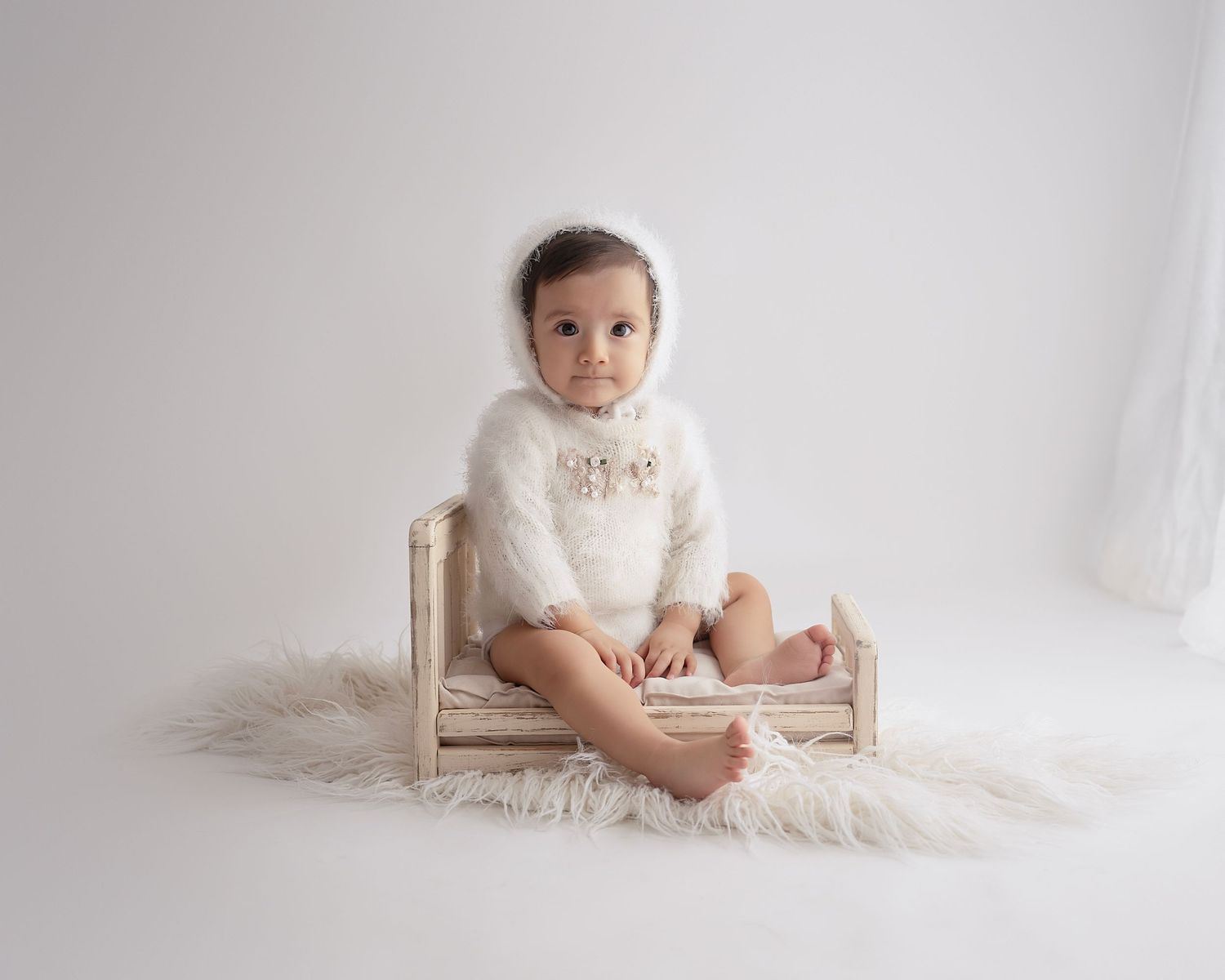 Small child wearing white fuzzy hoodie sitting on miniature wooden bed with fur rug.
