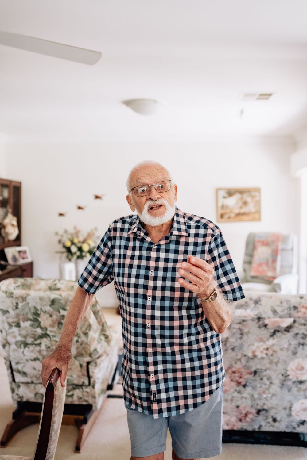An elderly man stands, leaning on a dining chair, seemingly telling a story.