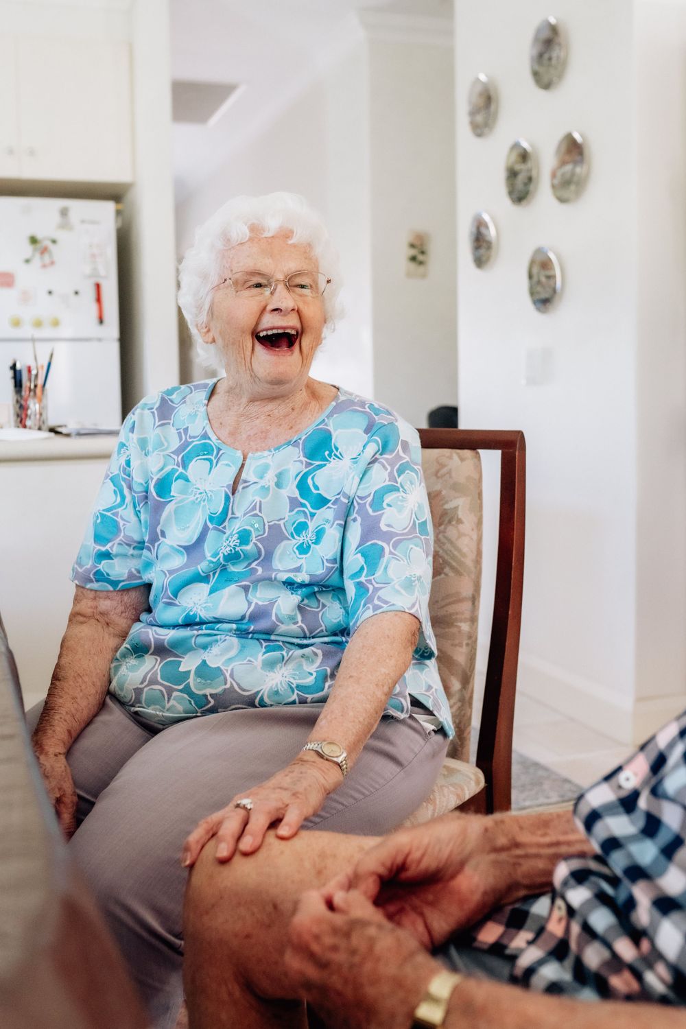 An elderly woman sitting on a chair, with a hand placed on an elderly man’s leg, laughing.