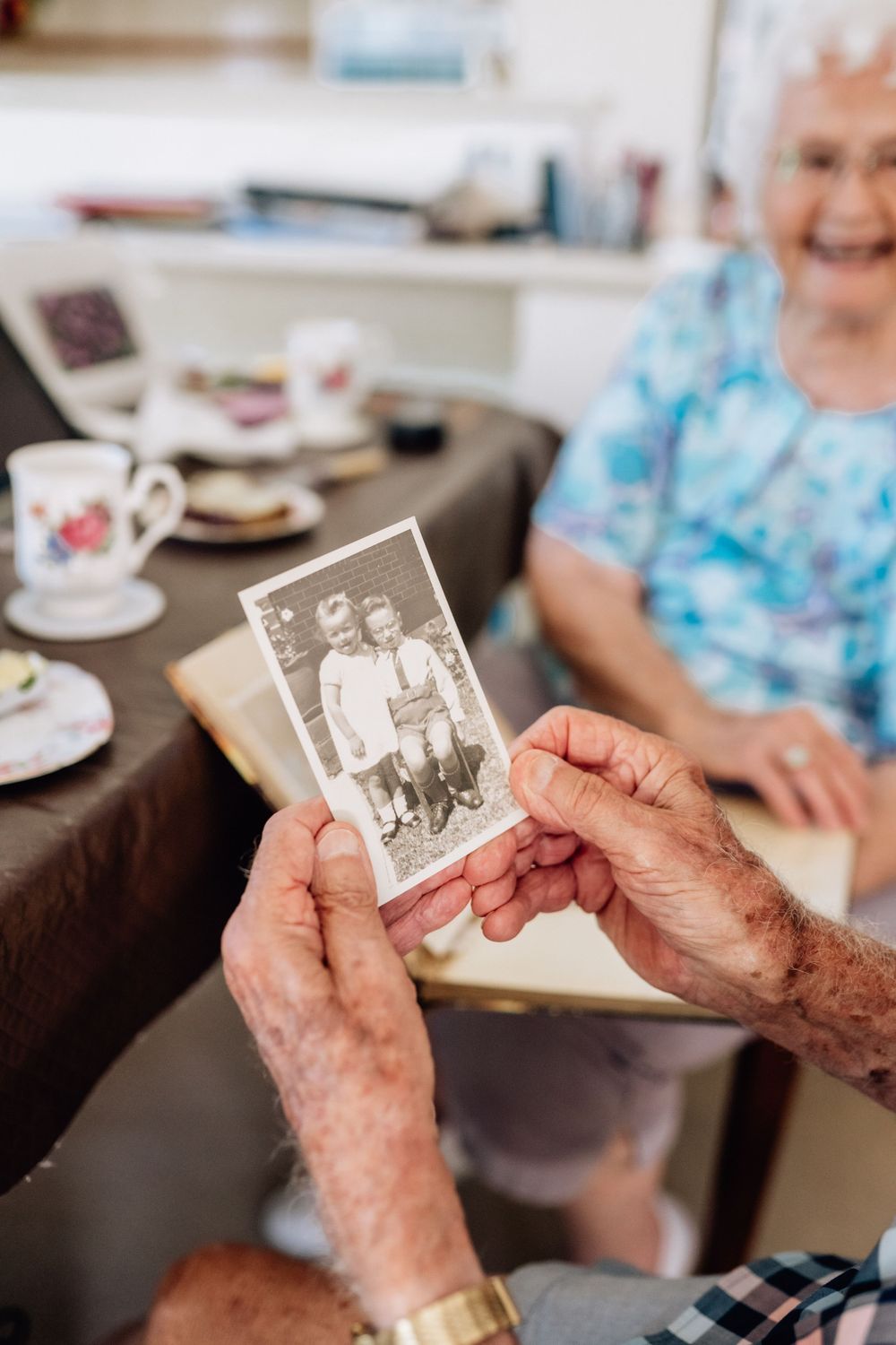 An elderly man holds an old photos of two young children, with an elderly women out of focus in the background of the image.