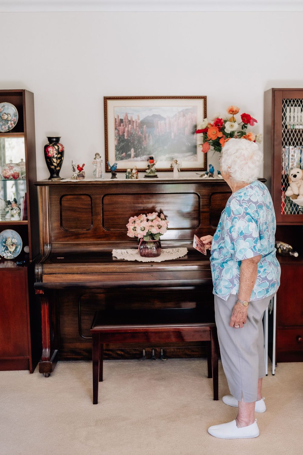 An elderly woman stands in front of a brown upright piano, looking at a framed photograph on the wall above it.