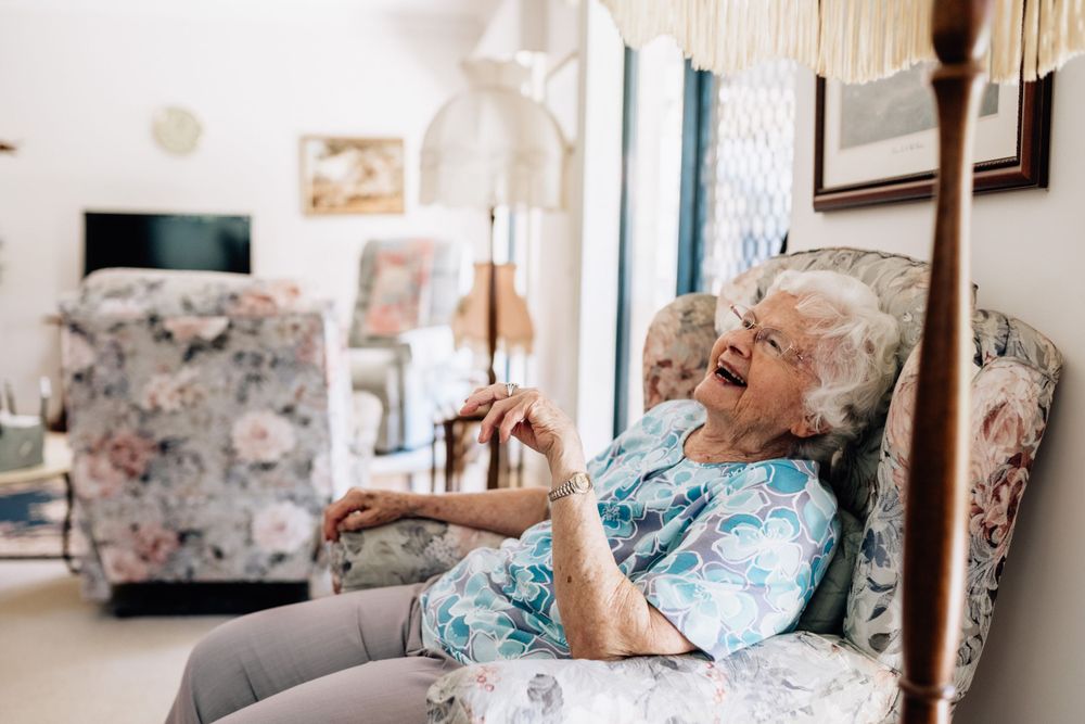 An elderly woman reclines in an arm chair, laughing.