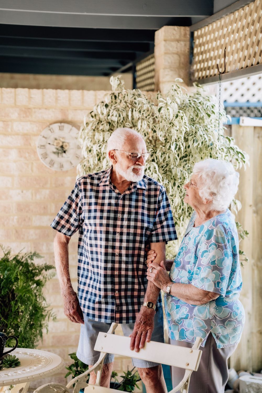 An elderly man and woman stand behind a garden bench, looking at each other and smiling.