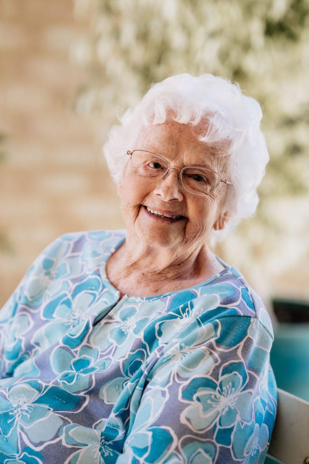 A portrait of an elderly woman, looking directly at the camera, seated in an chair outside.
