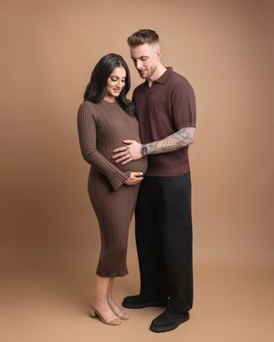 Couple in matching brown outfits posing for maternity photo, man embracing pregnant woman against tan background.
