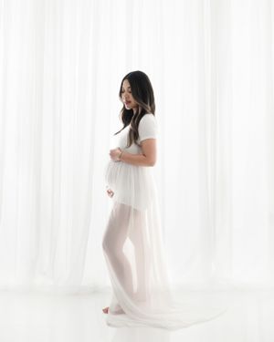 Ethereal maternity session with flowing white sheer dress in bright studio.