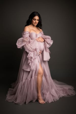 Dreamy maternity boudoir in flowing mauve tulle gown against dark backdrop.