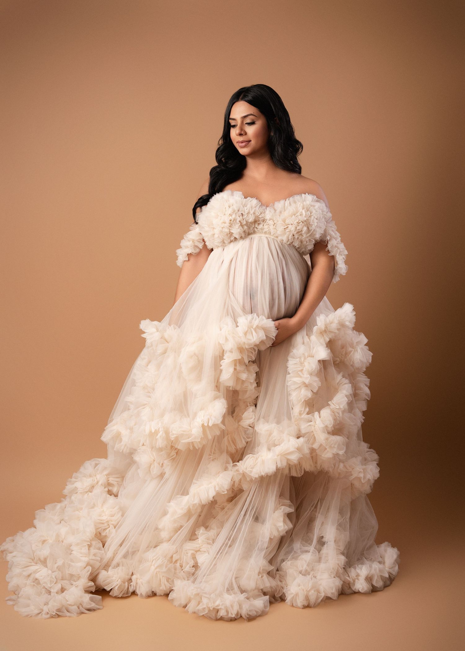 Dreamy maternity photos in flowing white ruffled gown against warm brown background.
