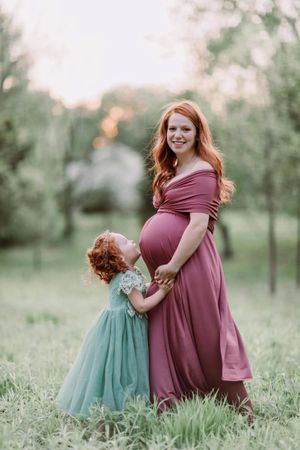 Sweet outdoor maternity moment with child in turquoise dress amid spring greenery.