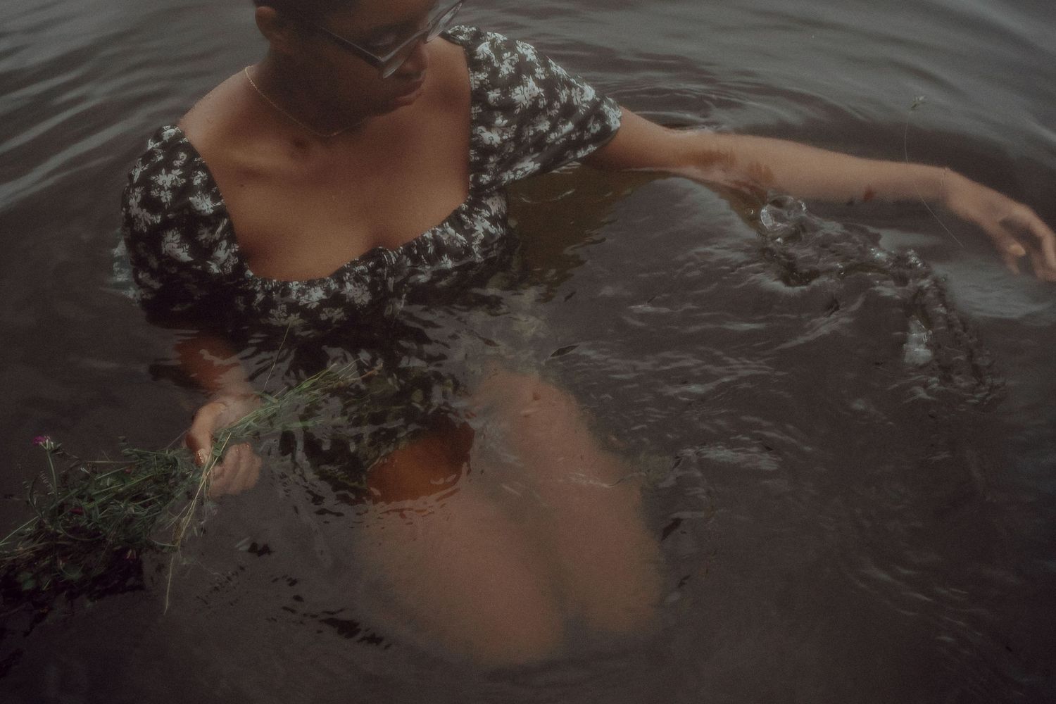 Figure in a floral dress wades through dark rippling water in moody lighting.