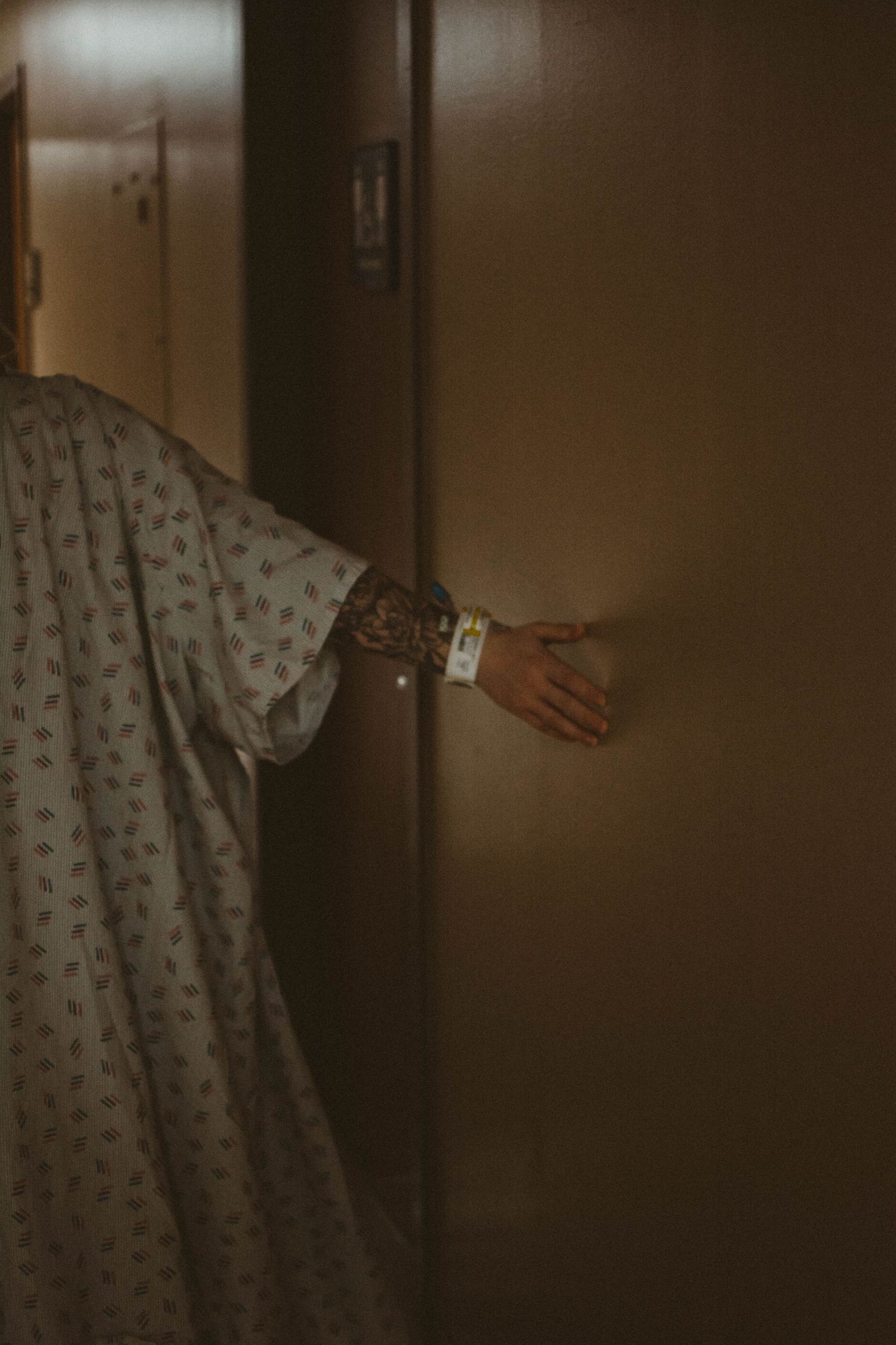 Patient in hospital gown reaching for door handle in dimly lit hallway.