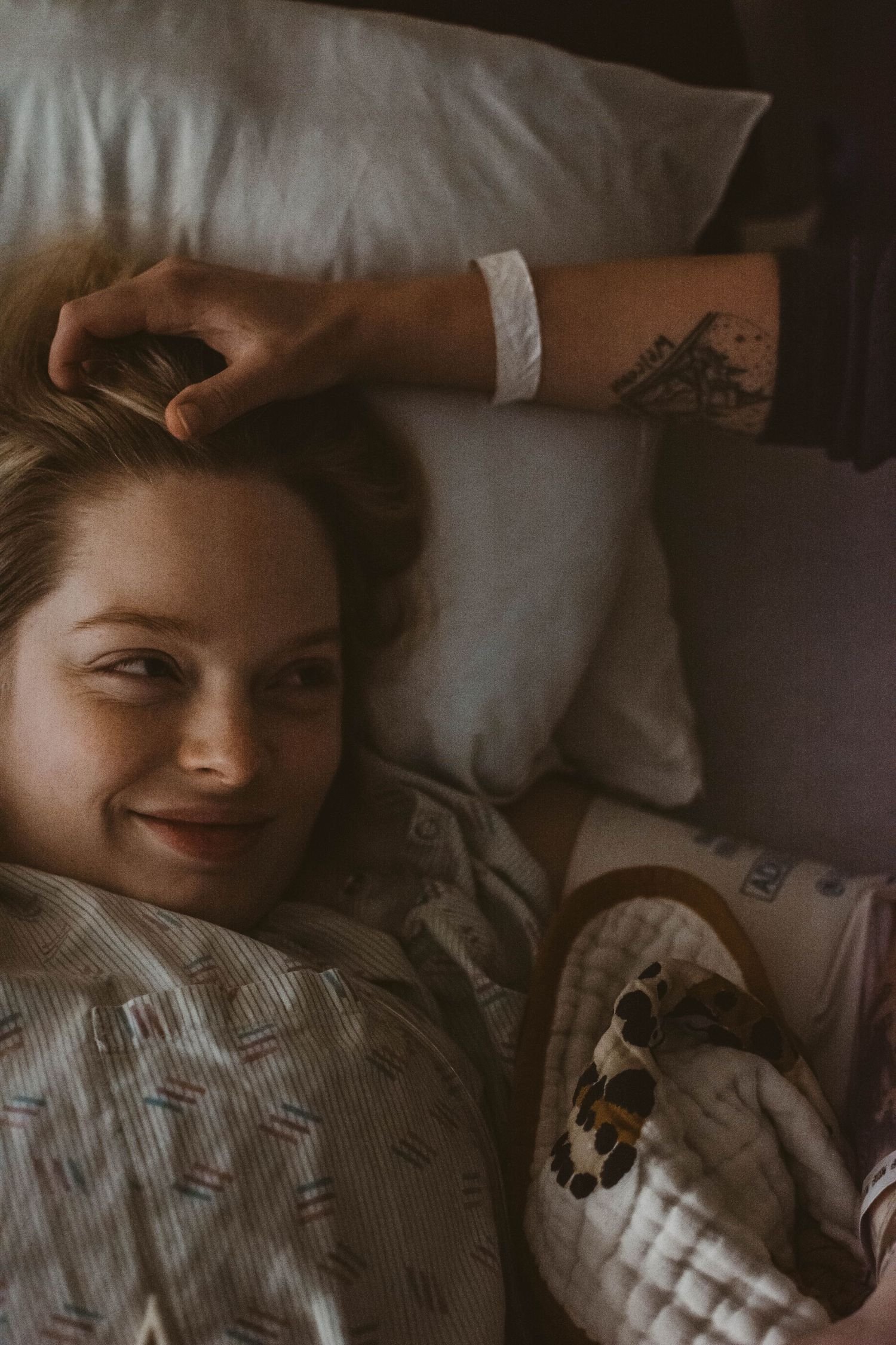 Person with tattoo and hospital bracelet resting on white pillows with gentle smile.