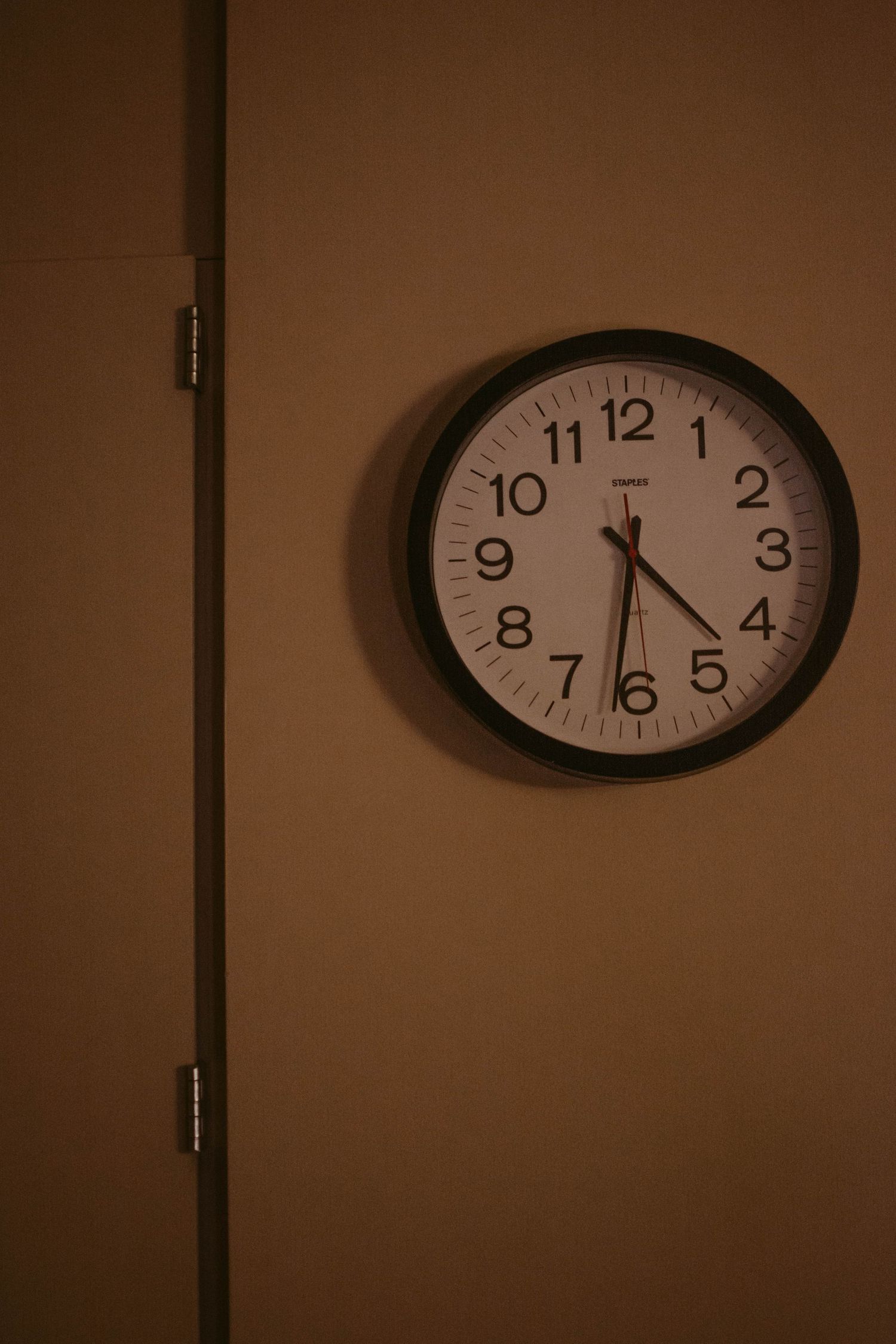 Wall clock showing 4:25 against beige wall in low light.