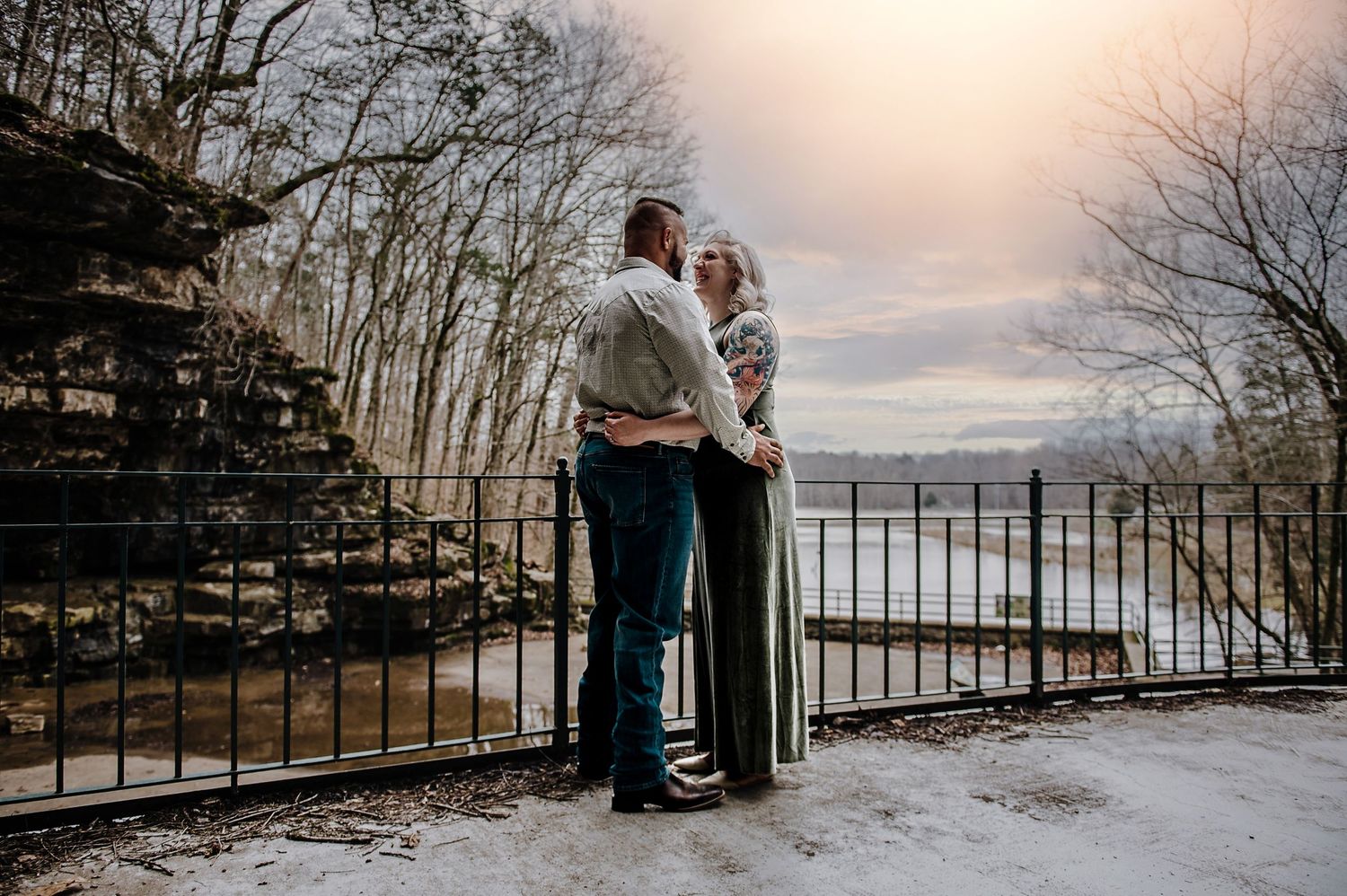 A couple stands close together on a scenic overlook during winter with bare trees in the background.