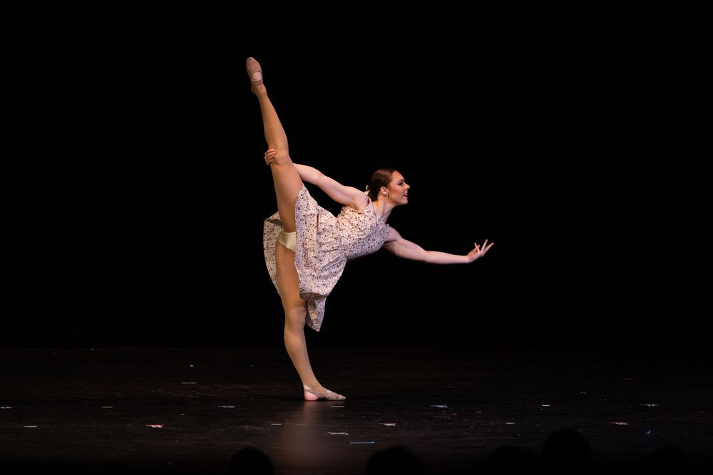 A ballet dancer performs an elegant arabesque pose in a light colored costume on a dark stage during a performance.