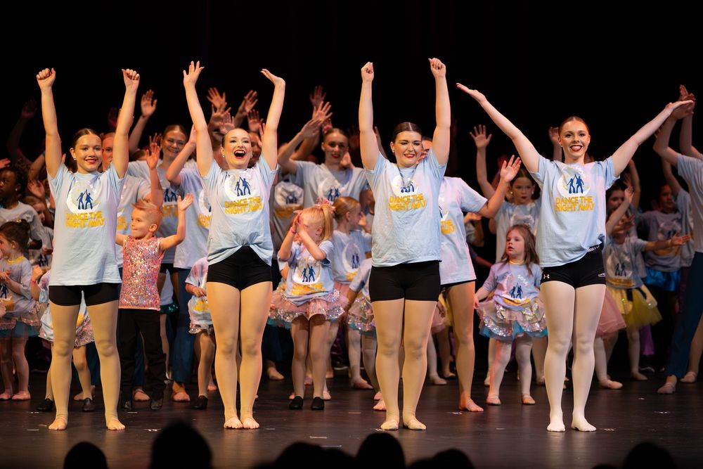 Dance students in matching white t-shirts and black shorts perform on stage with their arms raised during a recital.