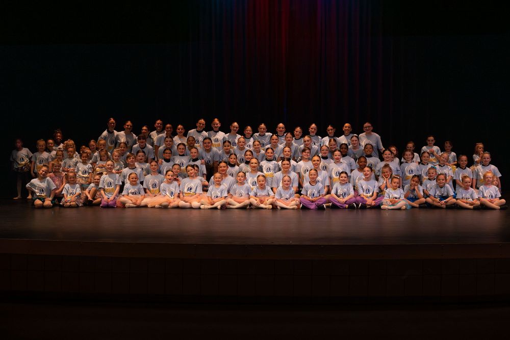 Large group of dancers in white costumes posing together on stage for a performance photograph.