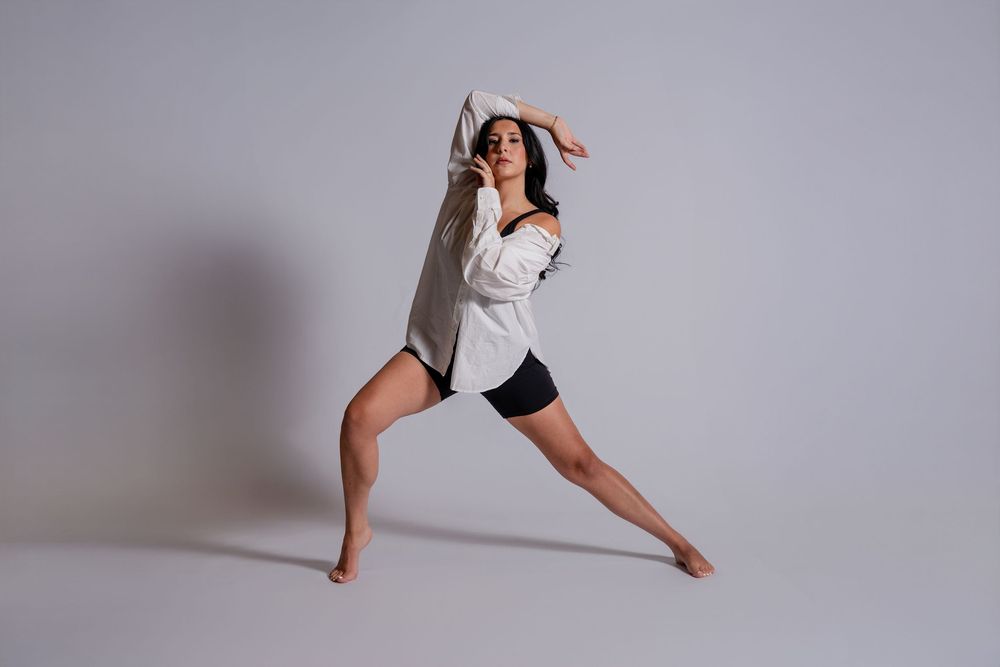 Dancer in white top and black shorts strikes an elegant lunging pose against a gray studio backdrop while extending arms gracefully.