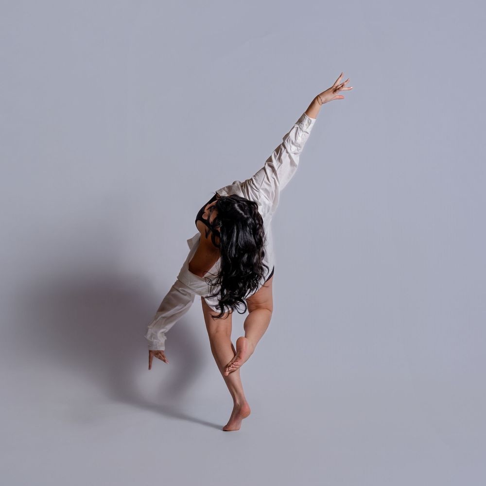 A dancer in white attire performs a graceful contemporary dance movement against a gray studio backdrop.