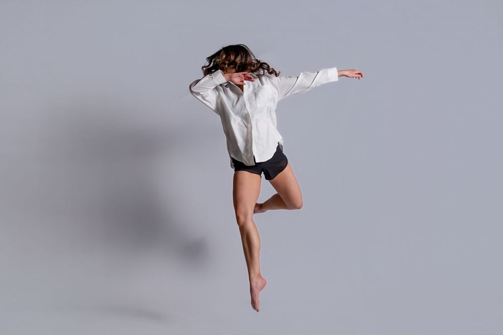 A dancer in a white shirt and black shorts leaps gracefully through the air against a gray background.
