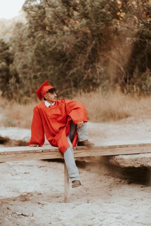 Graduate in red regalia reclines on wooden bench for artistic portrait.