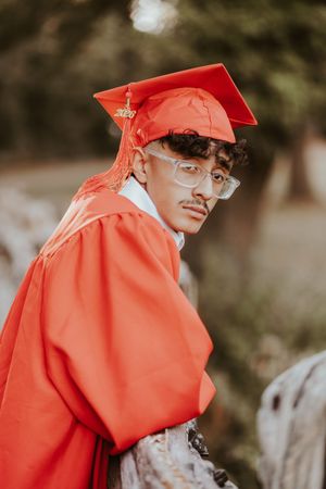 Graduate wearing bright red academic regalia and glasses poses against a blurred outdoor background.