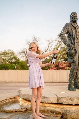 Person in light dress poses playfully near bronze statue outdoors.