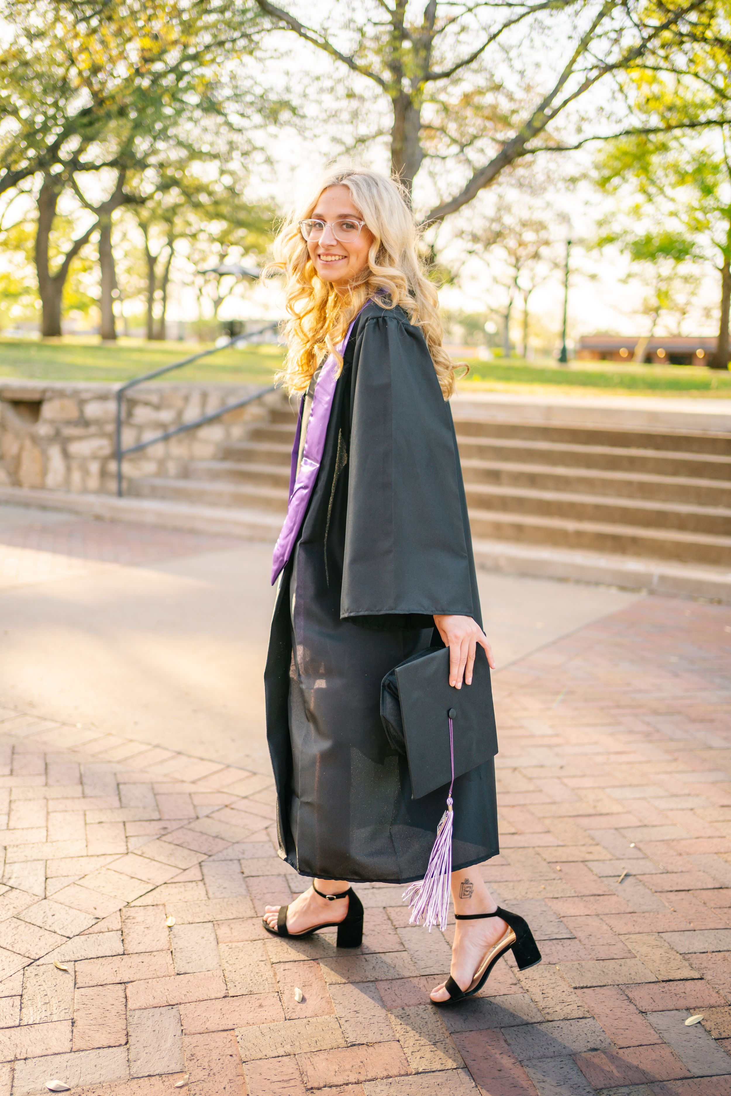 Graduate in black gown poses on brick pathway during sunset celebration.