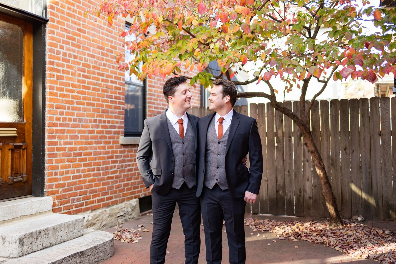 Grooms in dark suits laughing and chatting while walking together outside a brick building.