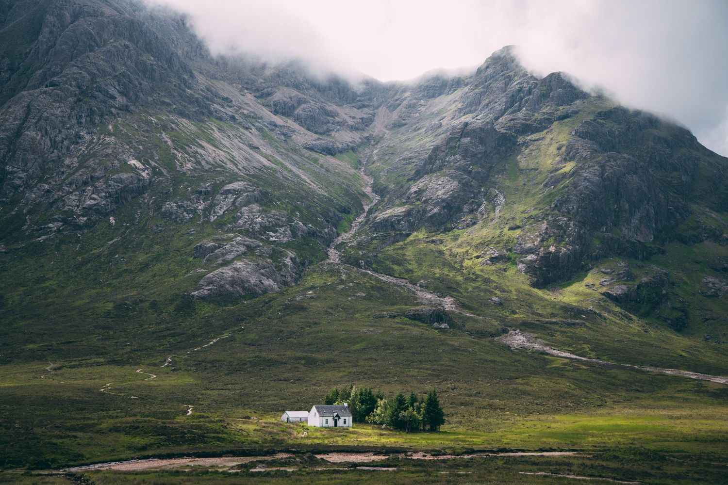 Lone white cottage surrounded by trees sits beneath towering mountain cliffs in misty Scottish landscape.