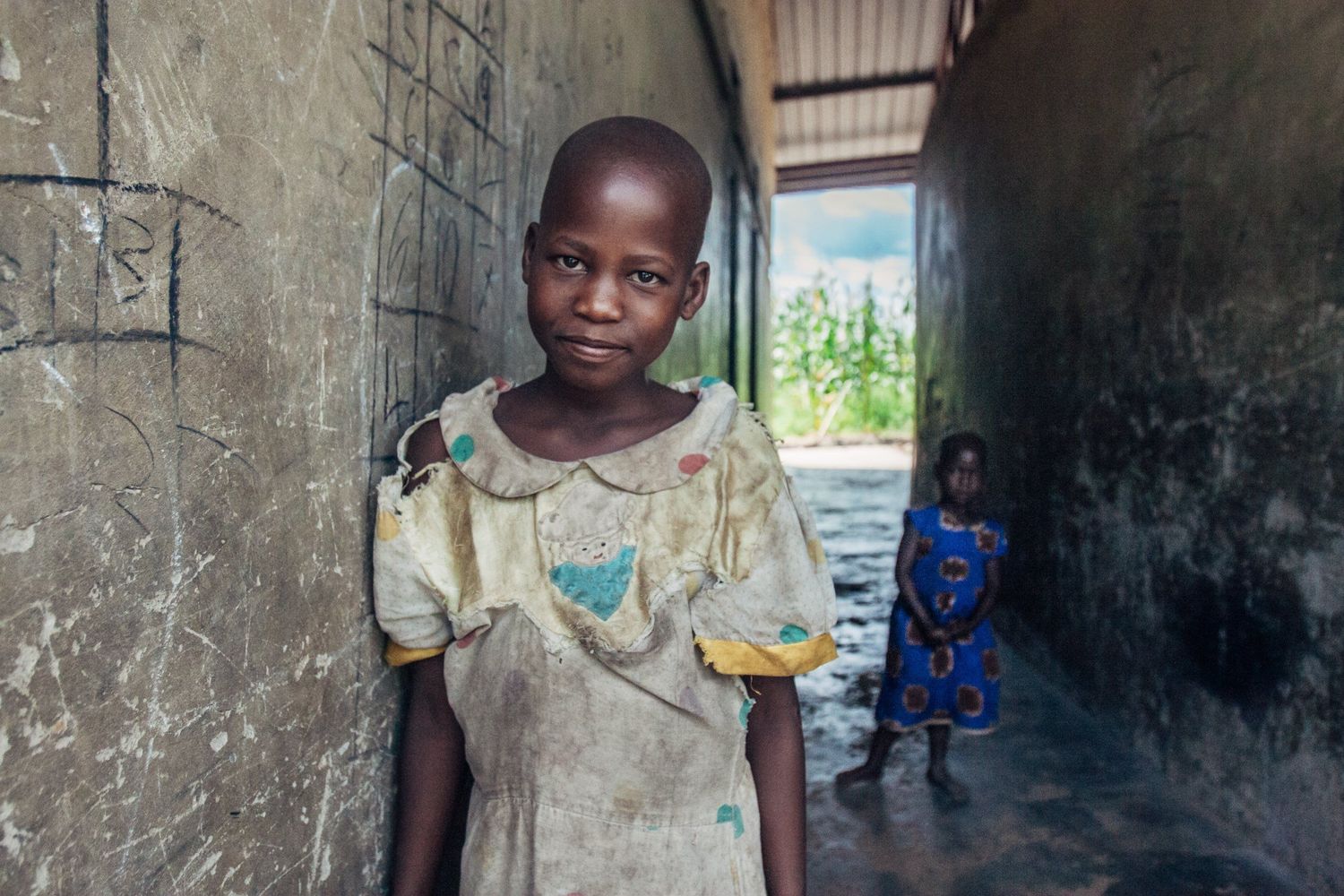 A small child stands in a worn concrete doorway wearing a tattered t-shirt.