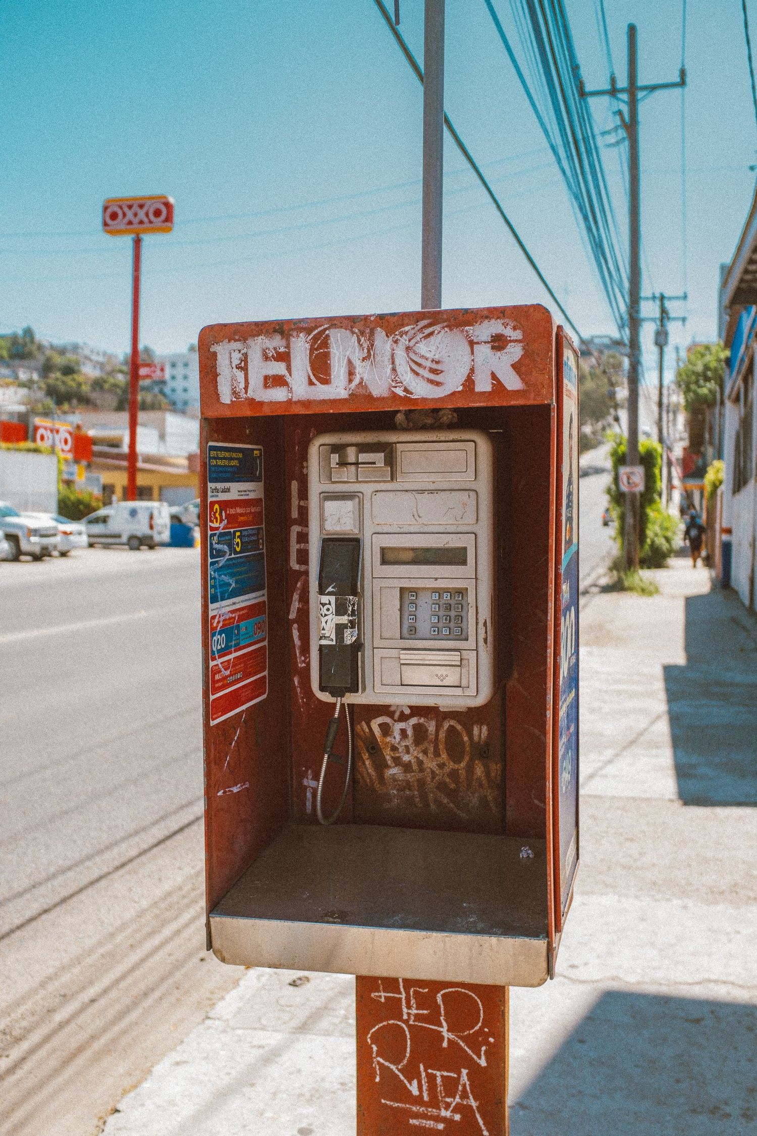 A vintage red telephone booth stands on a sunny street corner with power lines overhead.
