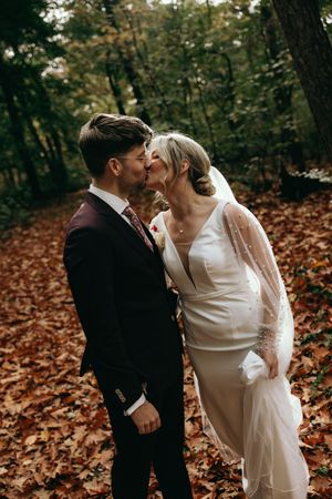 Romantic autumn wedding scene with couple kissing among fallen leaves in a forest setting.